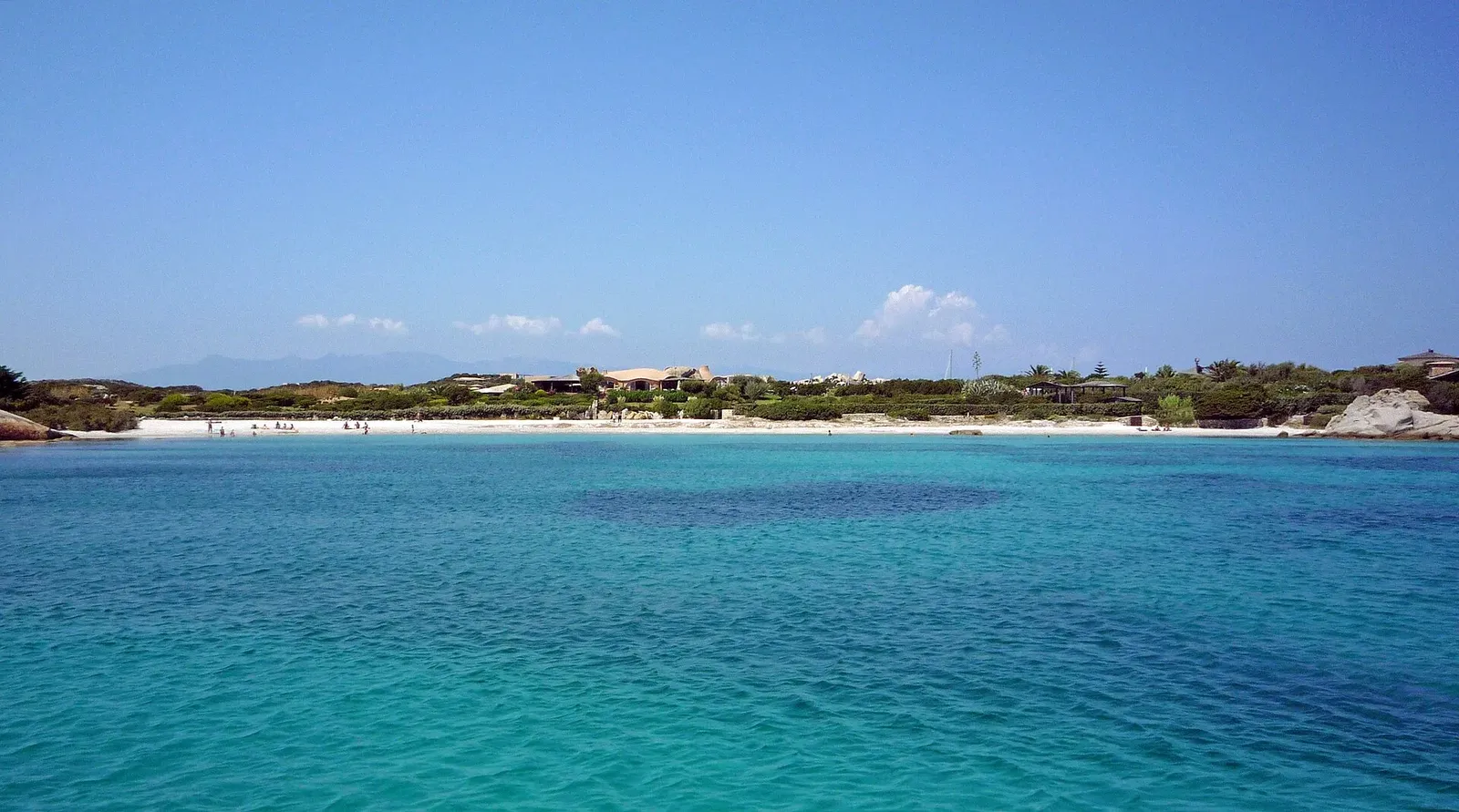 Eau turquoise devant une plage de sable blanc bordée de végétation et de villas basses, sur l'île de Cavallo