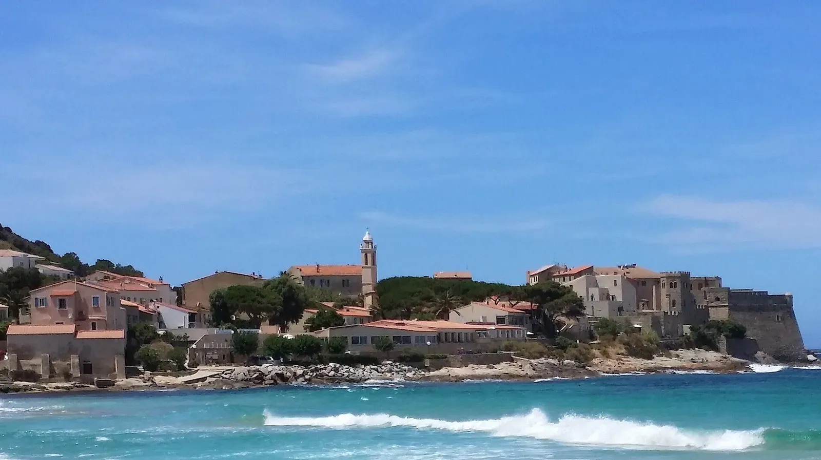 Maisons, clocher et citadelle d'Algajola au bord d'une mer turquoise, sous un ciel bleu, vus depuis la plage