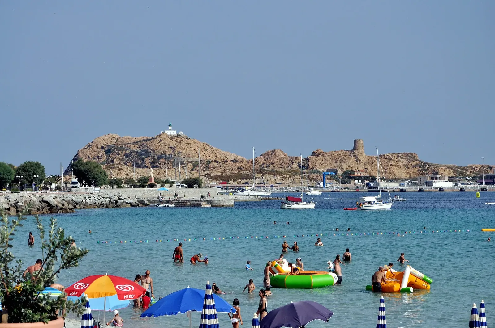 Baigneurs et parasols sur une plage aux eaux turquoise, avec un îlot rocheux portant une tour génoise et un phare en fond