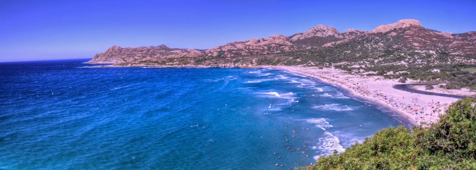 Vue panoramique d'une grande plage de sable bordée d'une mer bleu intense, avec des collines rocheuses et du maquis au fond