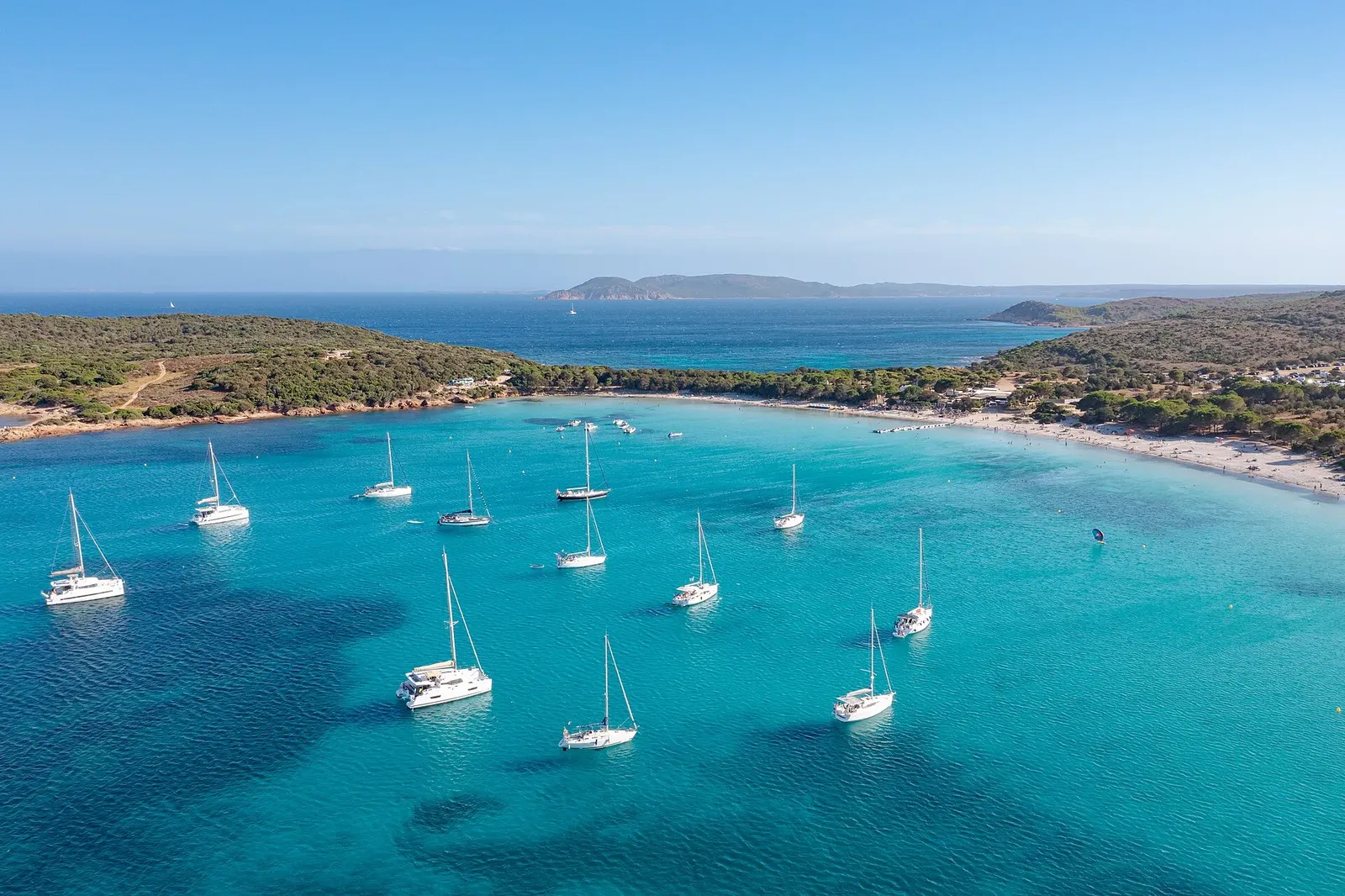 Vue aérienne d'une crique en arc de cercle à l'eau turquoise, avec des voiliers au mouillage et une plage de sable blanc.
